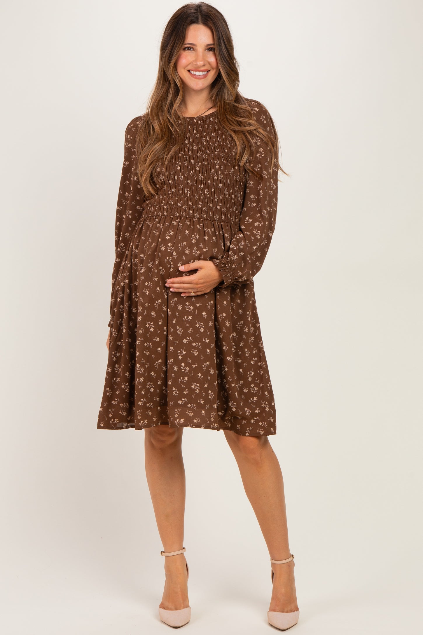 woman wearing a brown floral maternity dress with long sleeves, smiling and holding her baby bump