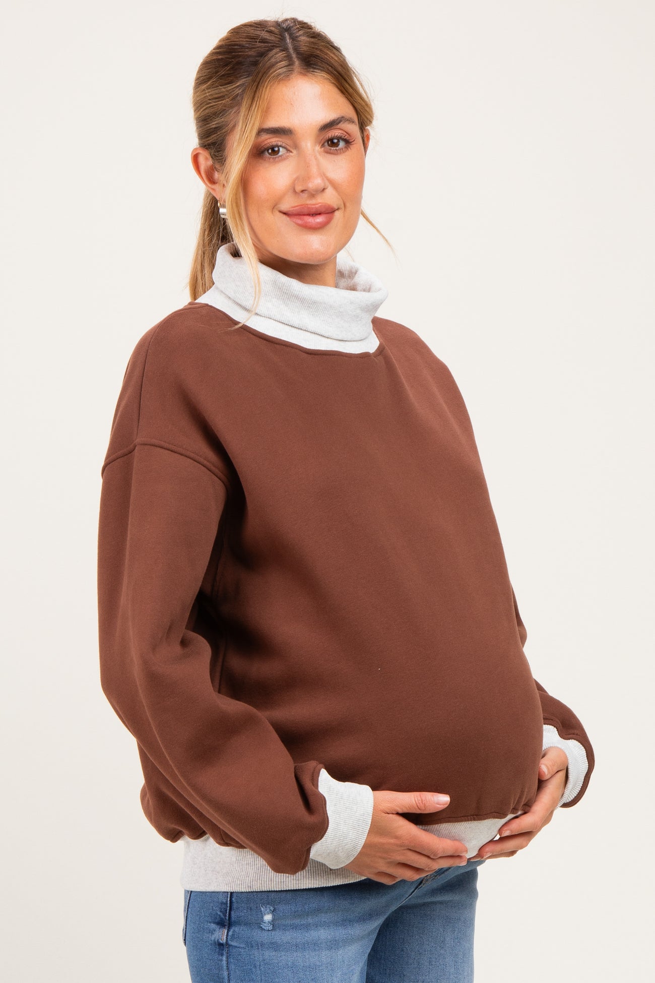 woman wearing a brown maternity sweatshirt with a gray turtleneck collar and holding her baby bump