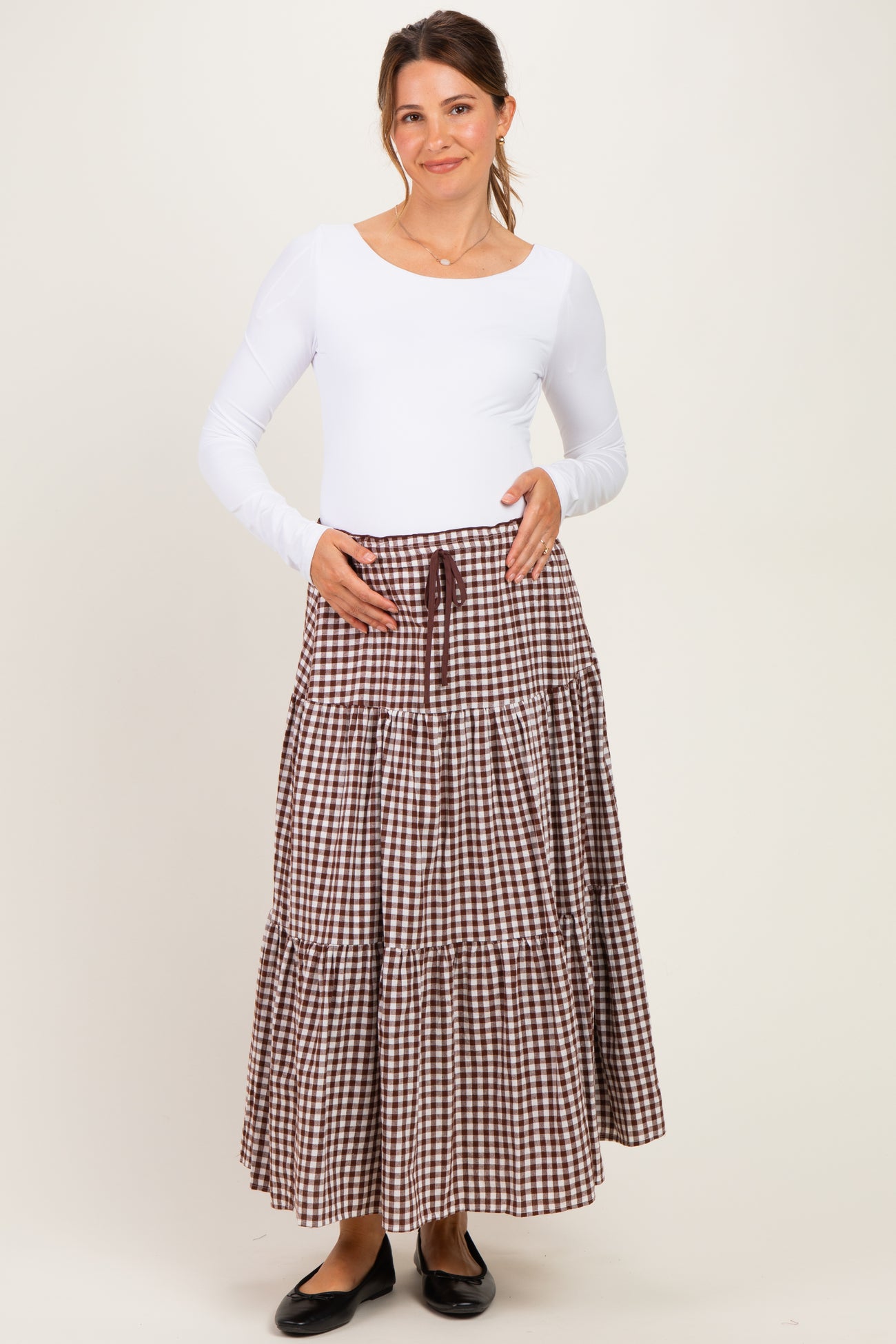 woman wearing a white long sleeve maternity top and a brown gingham tiered maternity skirt, holding her baby bump