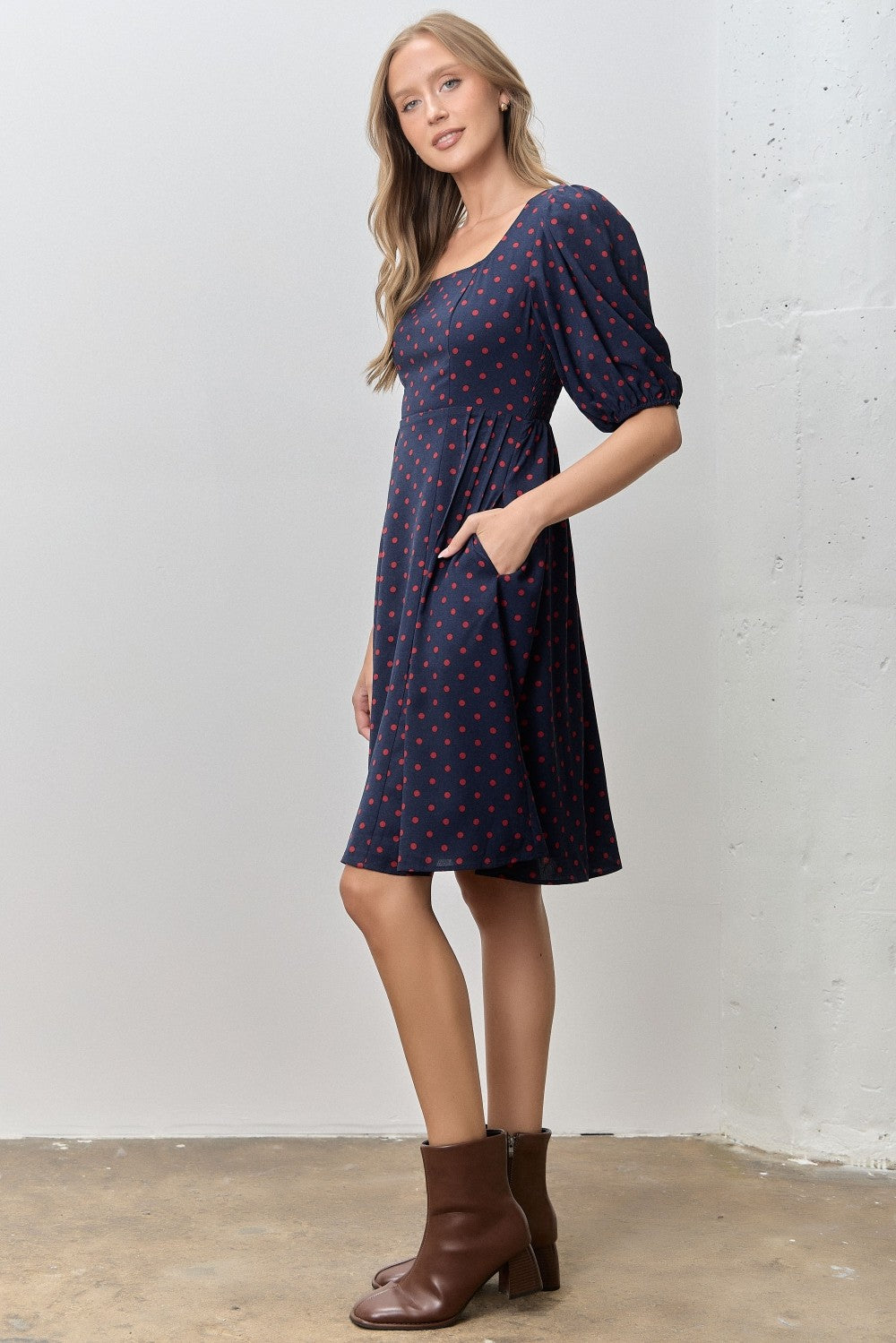 woman wearing a navy blue short sleeve dress with red polka dots and a square neckline, styled with brown ankle boots