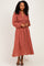 woman wearing a rust-colored long sleeve maxi dress with black polka dots and a smocked bodice, standing with hands on hips