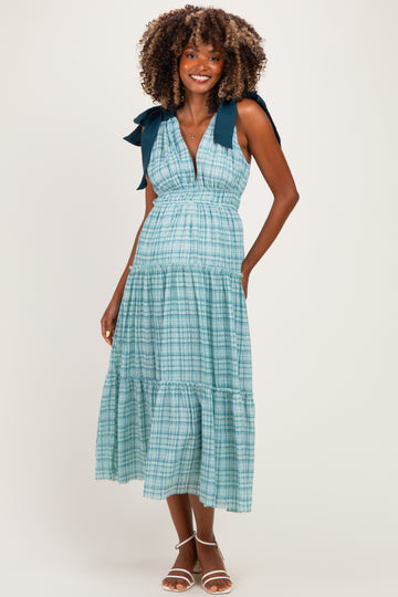 woman wearing a light blue plaid maternity dress with tie straps and a tiered skirt, showcasing her baby bump