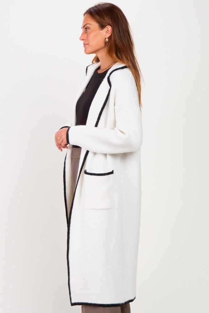 woman wearing a long white cardigan with black trim and pockets, standing sideways against a white background