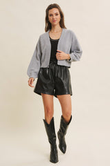 woman wearing a grey cropped cardigan, black tank top, and black leather high-waisted shorts, accessorized with tall black boots