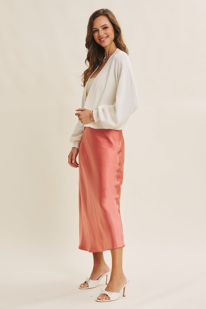 woman wearing a white oversized cardigan and a pink satin midi skirt with heels, smiling to the side