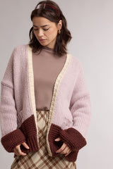 woman wearing a pink oversized cardigan with brown cuff details over a beige ribbed top and plaid skirt