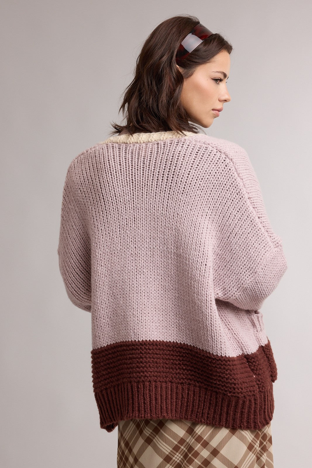 woman wearing a soft pink oversized knit sweater with a brown hem and checkered skirt, seen from the back