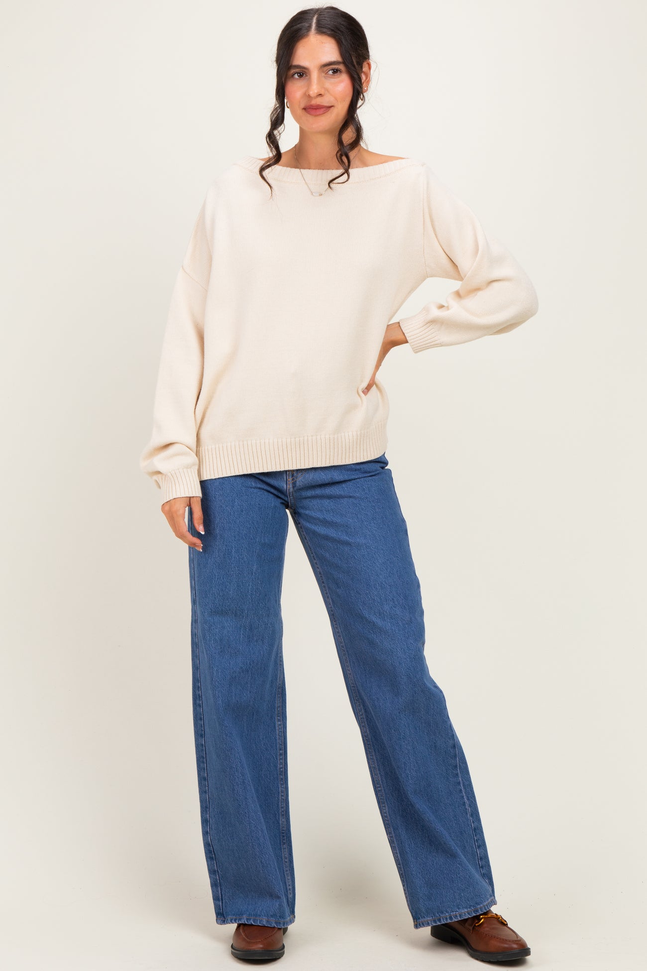 woman wearing a cream off-shoulder sweater and blue wide-leg jeans, standing against a light background