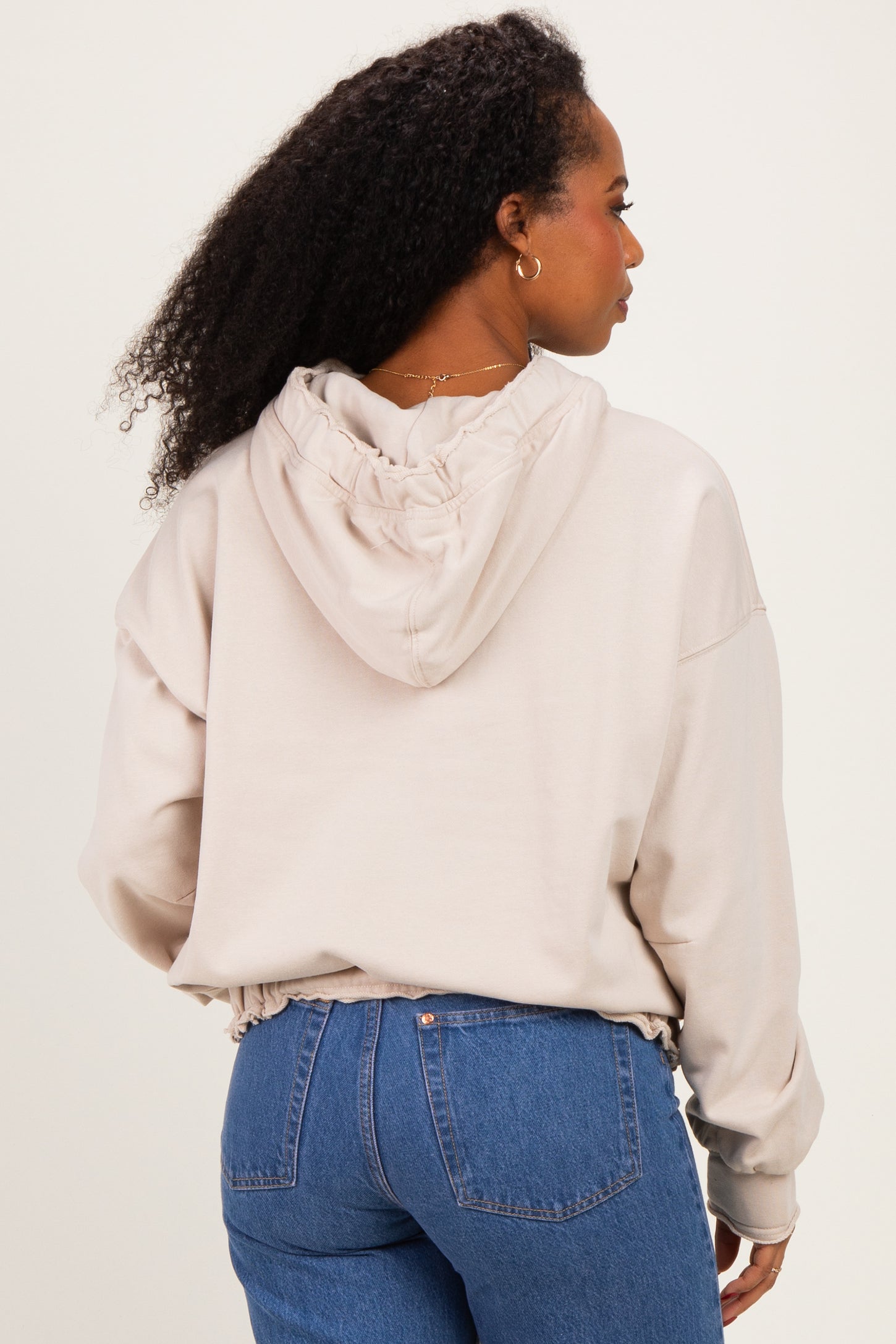 woman wearing a beige cropped hoodie with a ruched detail on the back and paired with high-waisted blue jeans