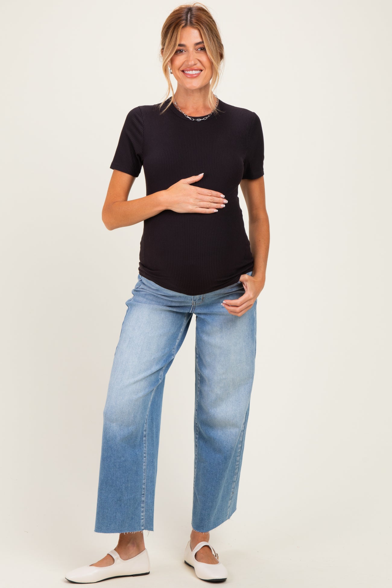 woman wearing a black ribbed maternity top and blue wide-leg jeans, holding her baby bump