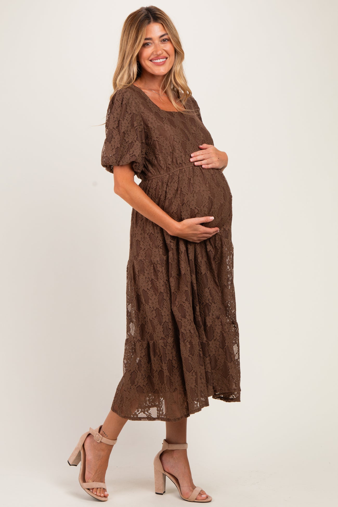 woman wearing a brown lace maternity dress and cradling her baby bump while standing on a neutral background