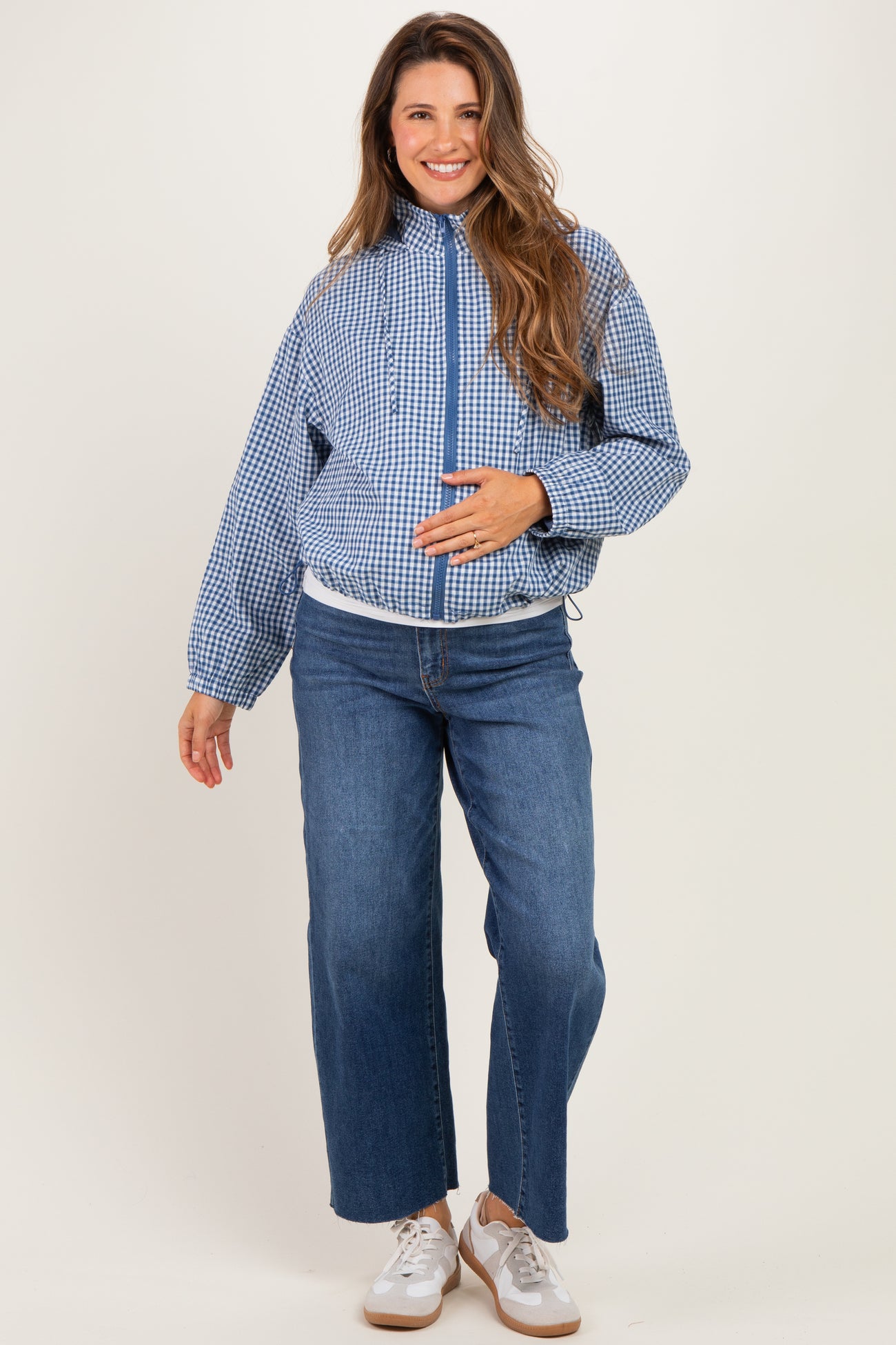 woman wearing a blue and white gingham maternity jacket and blue wide-leg jeans, gently cradling her baby bump