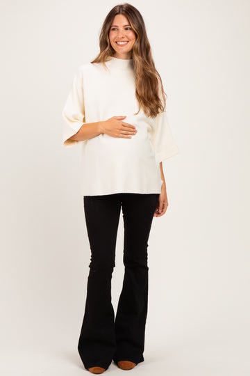 woman wearing a cream relaxed fit maternity top and black flared pants, gently holding her baby bump