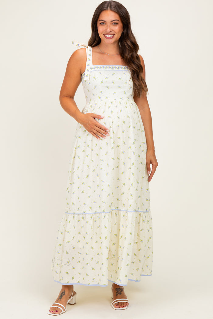 woman wearing a white floral maternity maxi dress with a square neckline and tying shoulder straps, gently cradling her baby bump