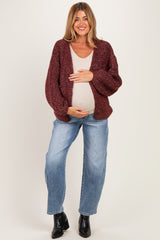 woman wearing a burgundy chunky knit maternity cardigan and light tank top, cradling her baby bump while standing in jeans