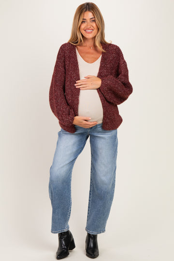 woman wearing a burgundy chunky knit maternity cardigan and light tank top, cradling her baby bump while standing in jeans