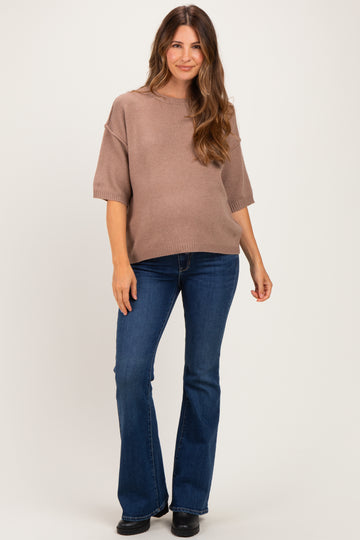 woman wearing a light brown short sleeve maternity sweater and blue flare jeans, gently cradling her baby bump