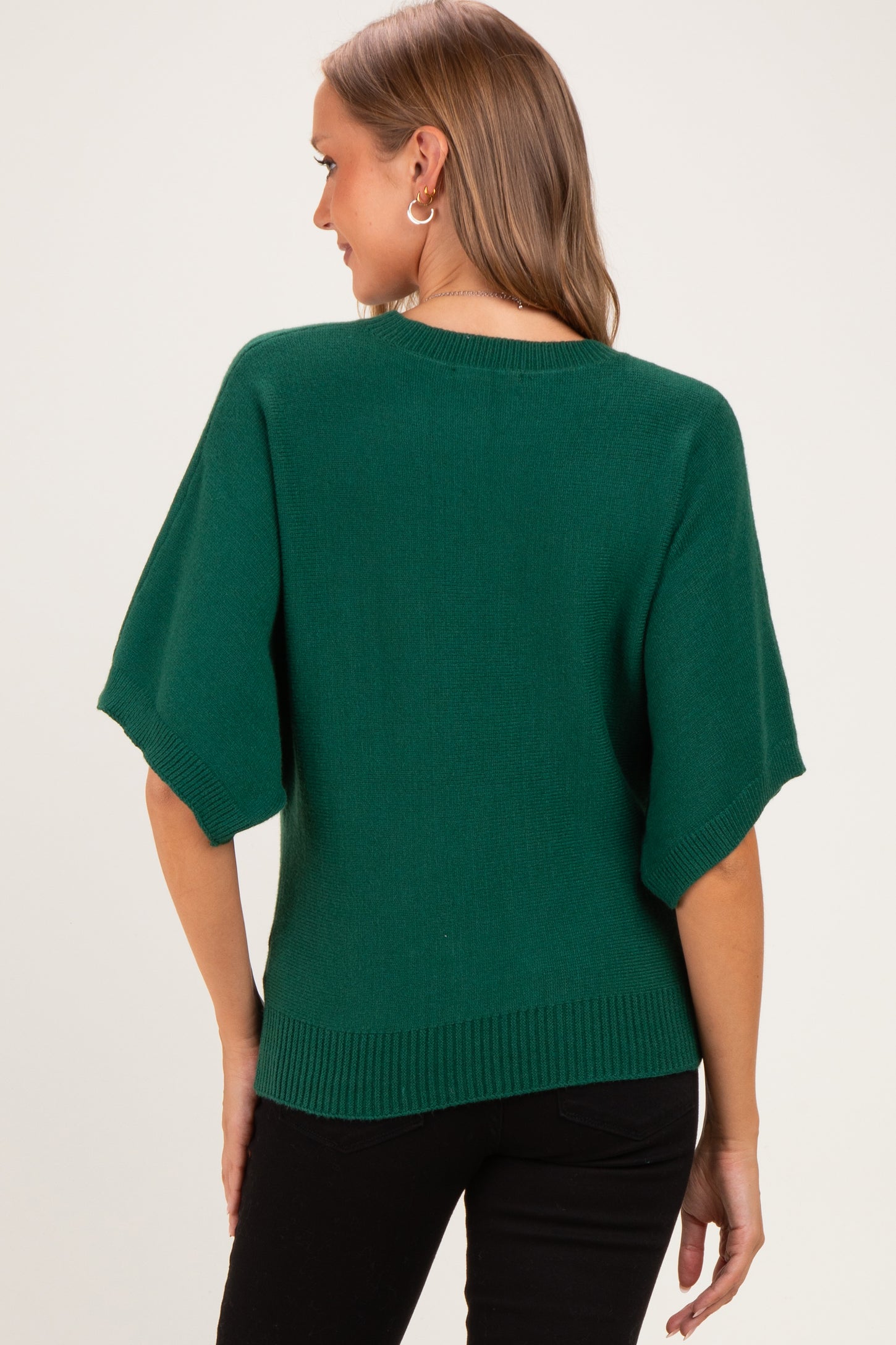 woman wearing a green short sleeve maternity sweater, view from the back, styled with black pants