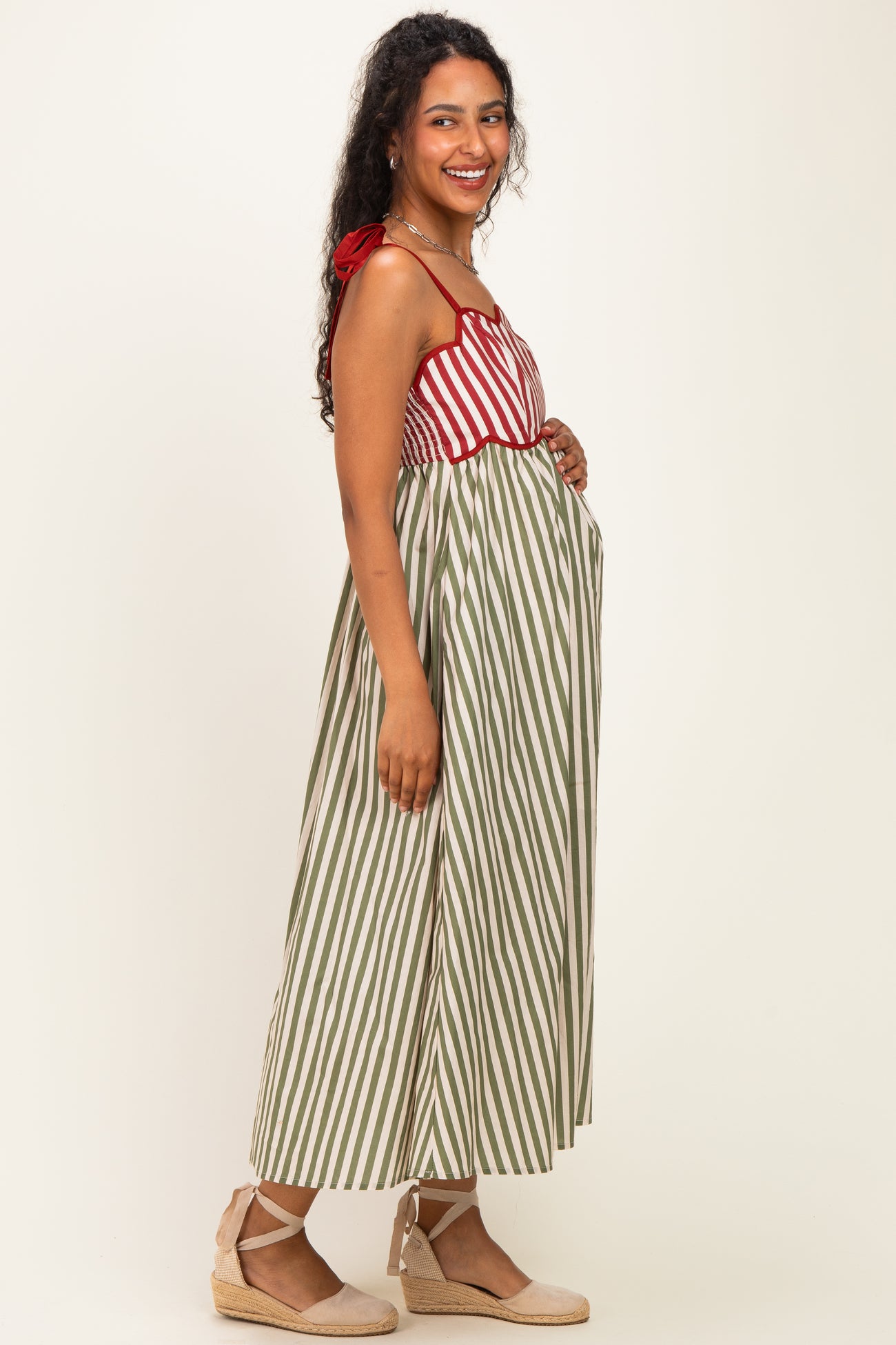 woman wearing a striped red and green maternity dress, smiling and showing her baby bump, with espadrille wedges