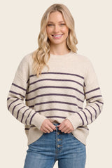 woman wearing a cream and purple striped knit sweater, smiling and holding her waist with blue jeans