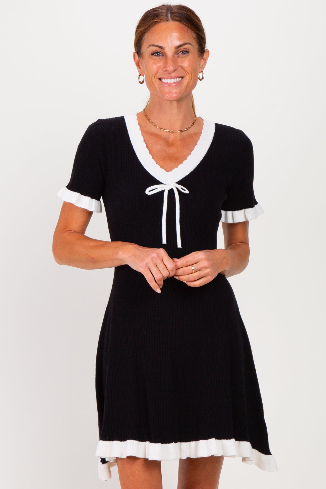 woman wearing a black short sleeve dress with white ruffle trim and a tie neckline standing and smiling