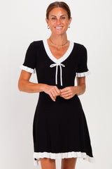 woman wearing a black short sleeve dress with white ruffle trim and a tie neckline standing and smiling