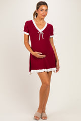 woman wearing a burgundy short sleeve maternity dress with a tie detail and white ruffled hem, holding her baby bump