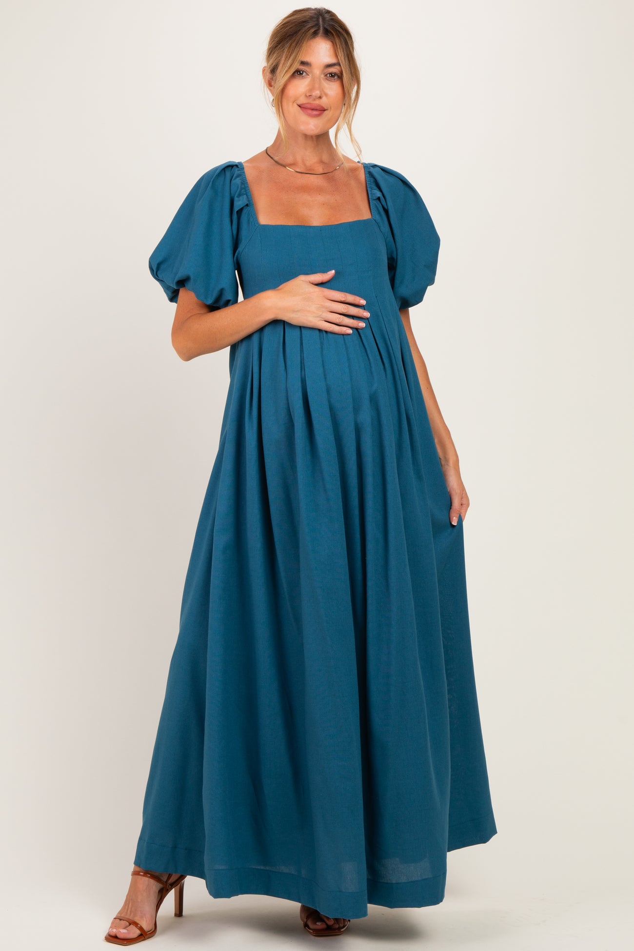 woman wearing a teal maternity maxi dress with puff sleeves and a square neckline, gently cradling her baby bump