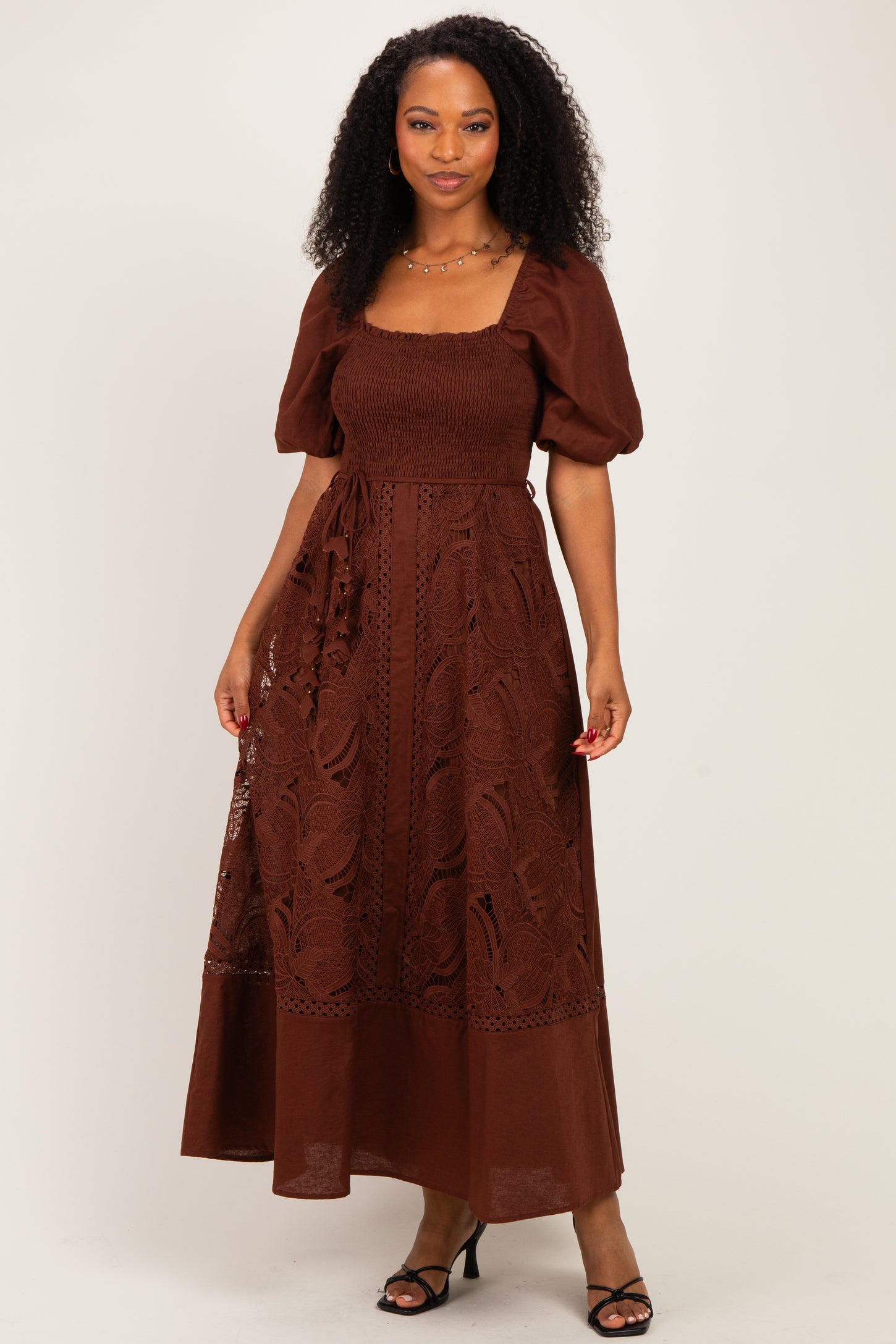 woman wearing a brown maxi dress with puff sleeves and lace detailing, standing with one hand on her hip