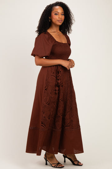 woman wearing a brown maxi dress with puff sleeves and embroidered details, standing against a neutral background