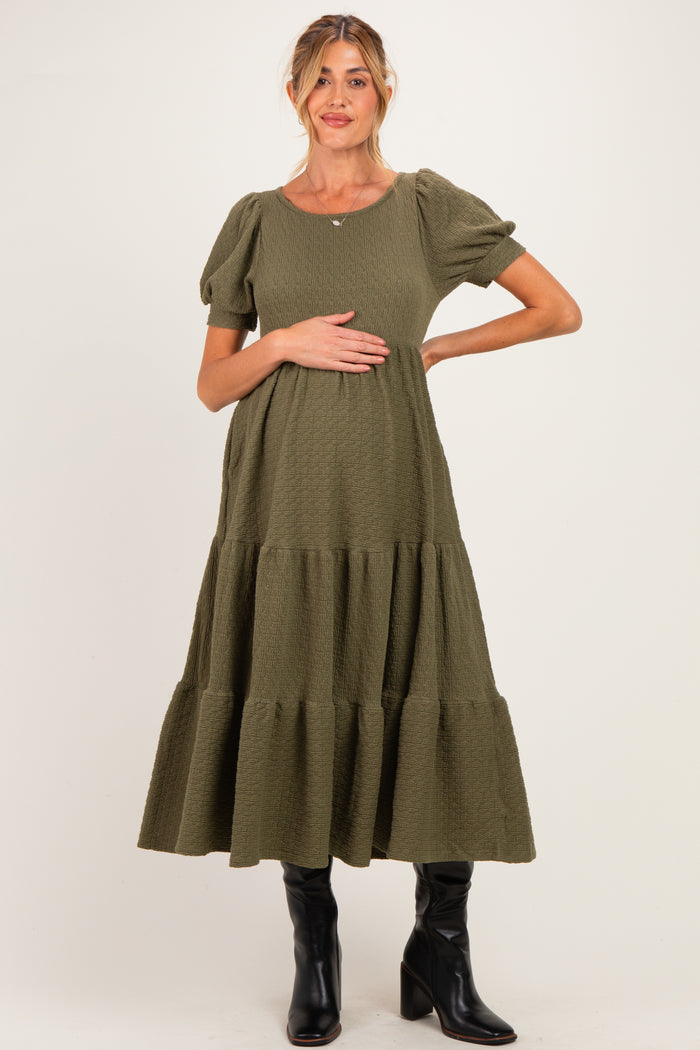 woman wearing an olive green maternity dress with puff sleeves and a tiered skirt, holding her baby bump