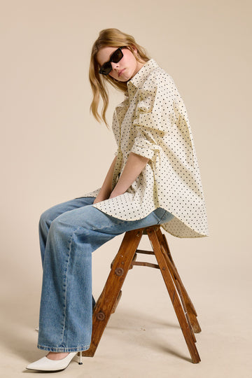 woman wearing a beige oversized shirt with black polka dots and high-waisted blue jeans, sitting on a wooden stool