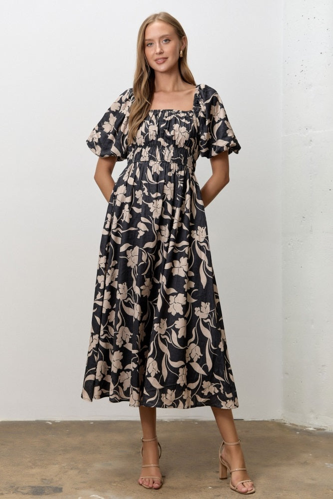 woman wearing a black floral midi dress with puff sleeves and a smocked bodice, standing with hands in pockets