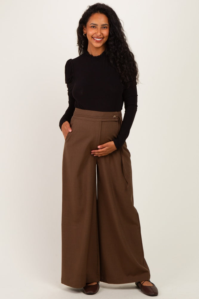 woman wearing a black long sleeve top and brown wide-leg maternity pants, holding her baby bump.