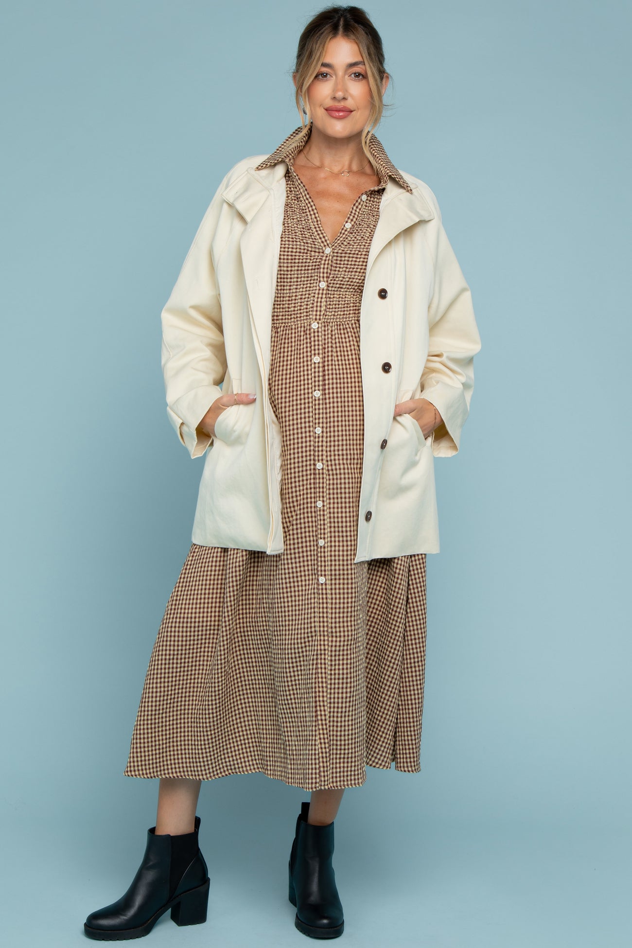 woman wearing a cream oversized jacket over a brown checkered maternity dress, with black ankle boots