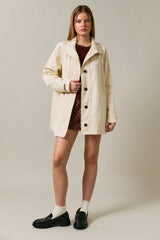 woman wearing a cream-colored button-up coat over a maroon top and plaid skirt, paired with black shoes and white socks