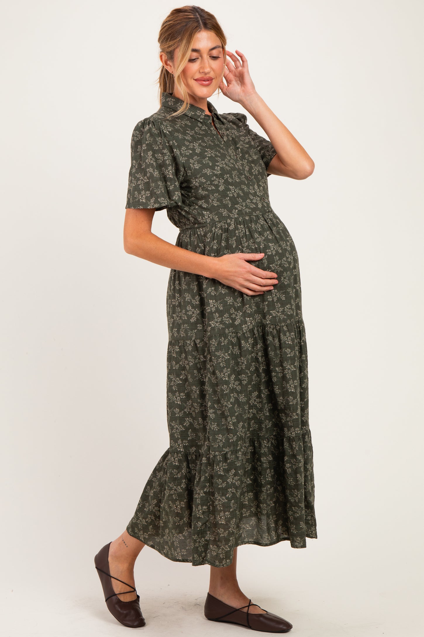woman wearing a green floral maternity dress with a tiered skirt, gently cradling her baby bump