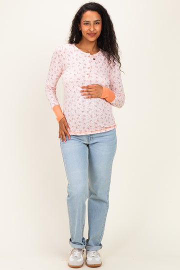 woman wearing a light pink floral maternity top with long sleeves and buttons, holding her baby bump and paired with light blue jeans