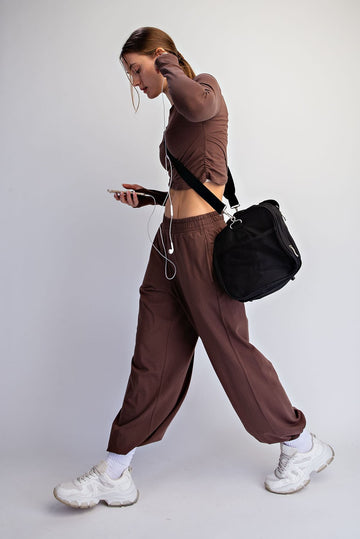woman wearing a brown long sleeve cut-out top and matching wide-leg pants, holding a phone with earphones and carrying a black bag