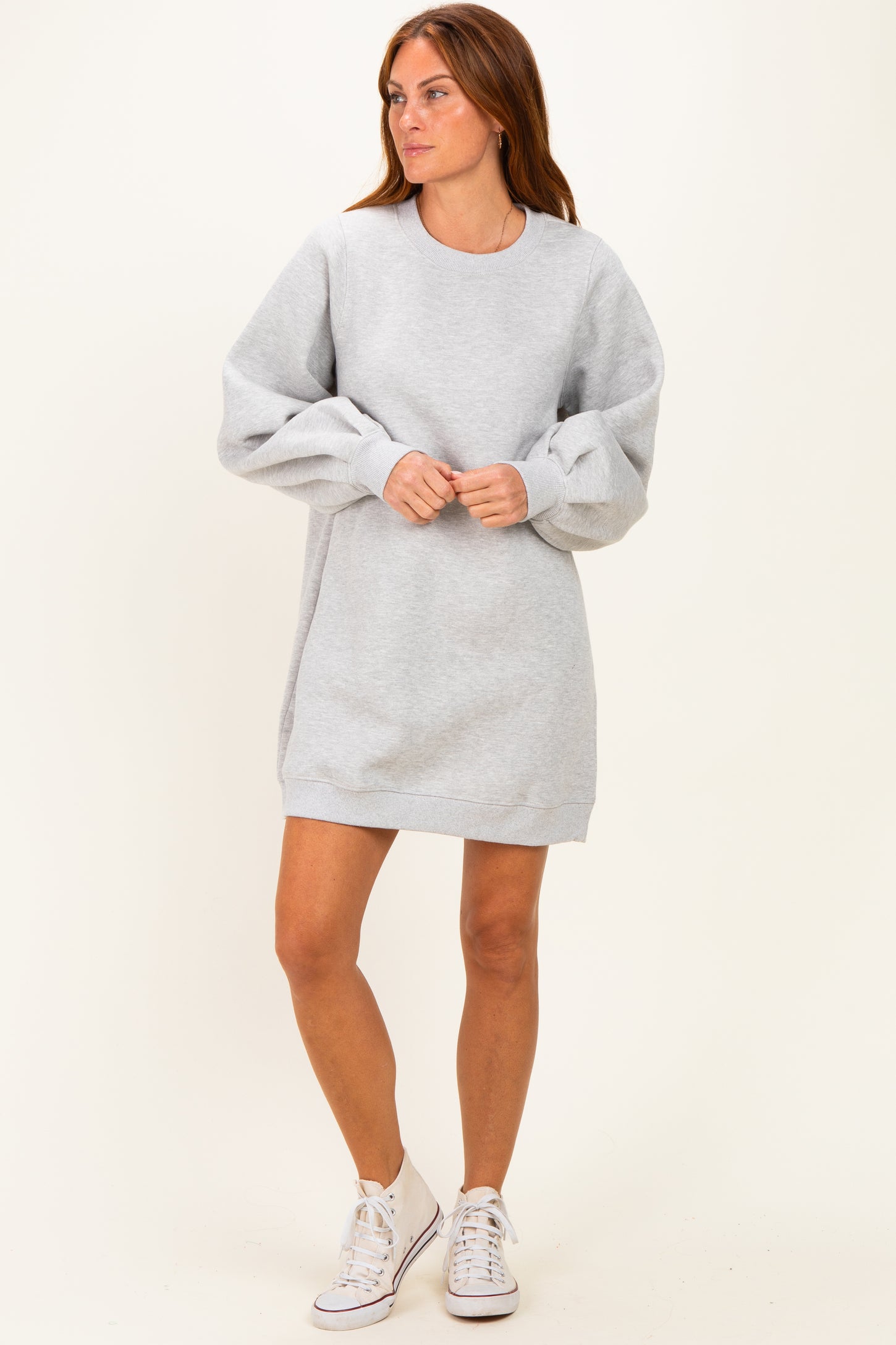woman wearing a gray sweatshirt dress with puff sleeves and white sneakers, standing against a neutral background