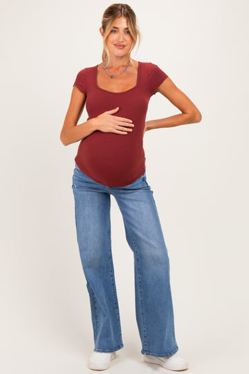woman wearing a burgundy short sleeve ribbed maternity top and blue flared jeans, cradling her baby bump