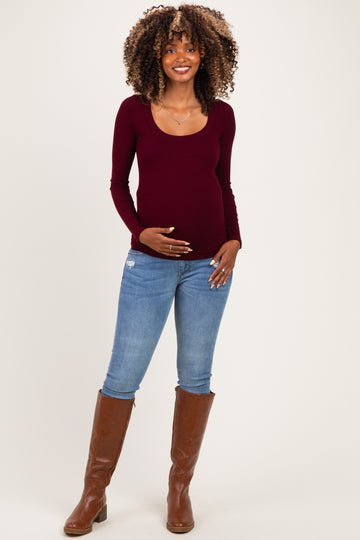 woman wearing a burgundy long sleeve maternity top and blue skinny jeans, gently holding her baby bump