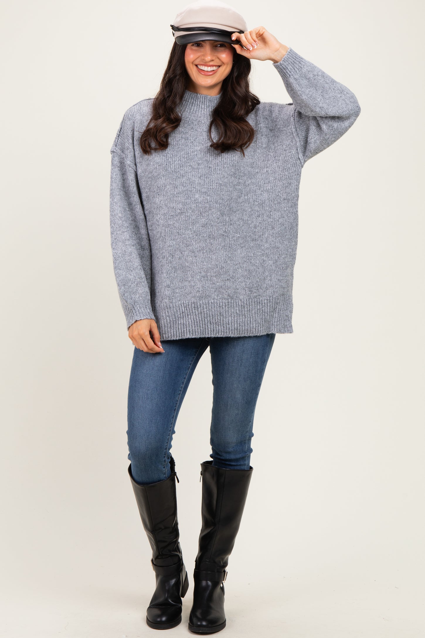 woman wearing a gray oversized sweater, blue skinny jeans, and tall black boots with a cap on her head