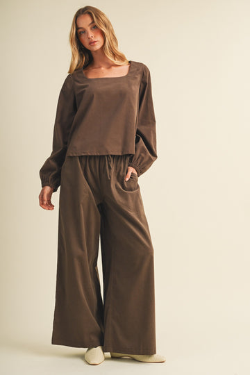 woman wearing a brown long sleeve top and matching wide-leg pants, posing with hands in pockets