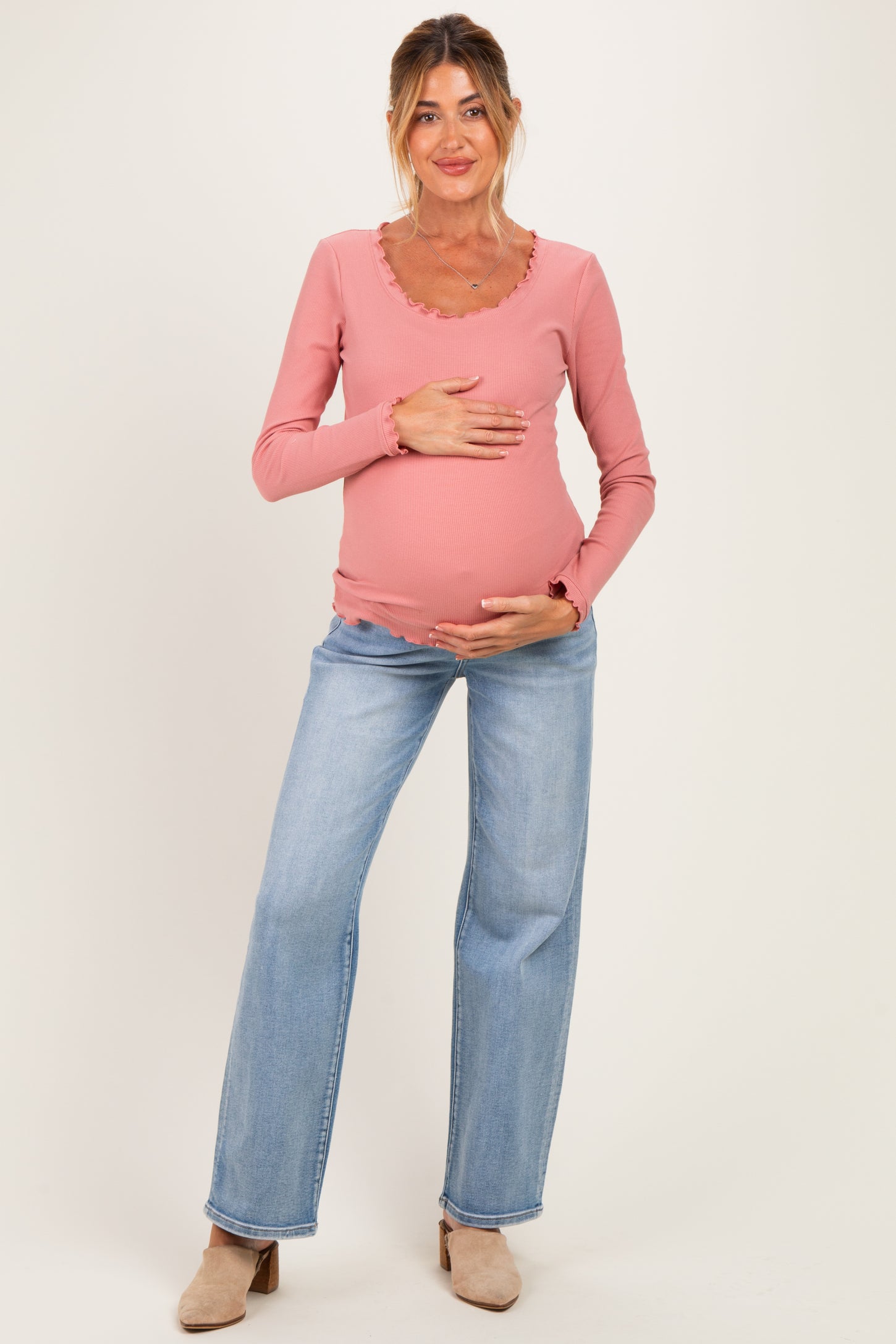 woman wearing a pink long sleeve maternity top with ruffled neckline and blue wide-leg jeans, holding her baby bump