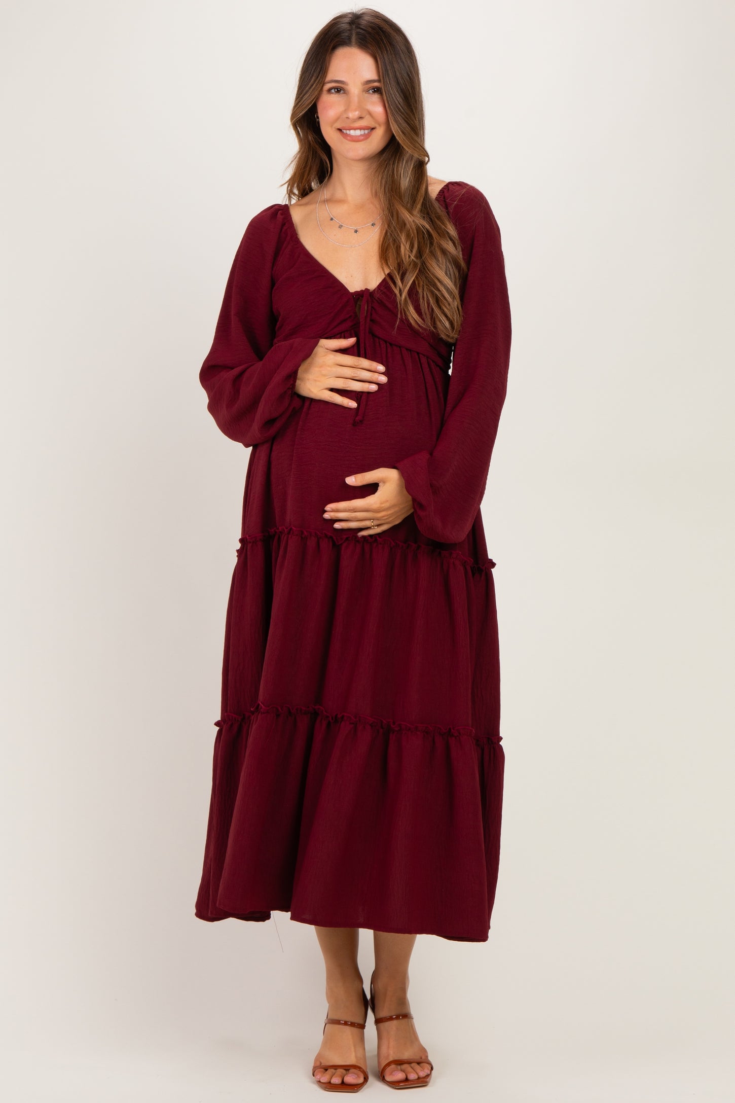woman wearing a burgundy maternity maxi dress with long sleeves and tiered ruffle details while holding her baby bump