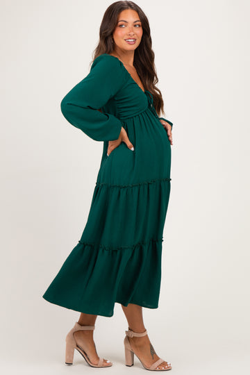 woman wearing a dark green maternity dress with a tiered skirt and long sleeves, posing with her baby bump visible