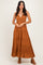 woman wearing a rust-colored maxi dress with a tiered design and eyelet details, standing with a slight pose