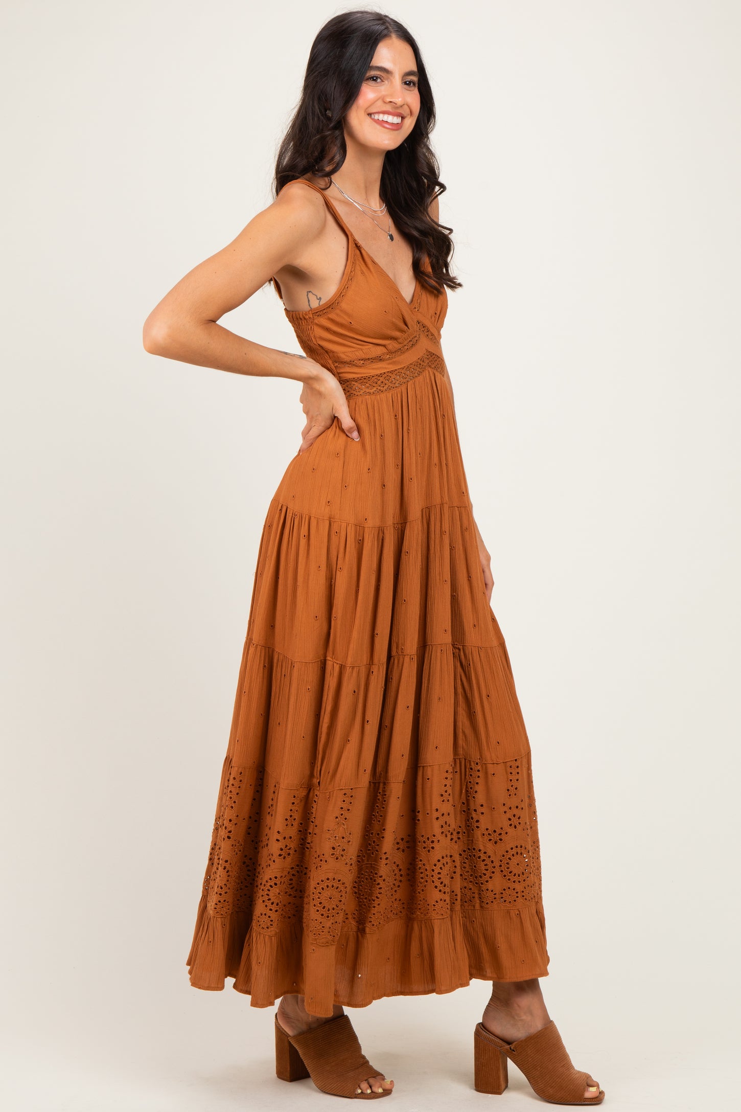 woman wearing a rust-colored maxi dress with a tiered design and eyelet details, paired with brown block-heel sandals