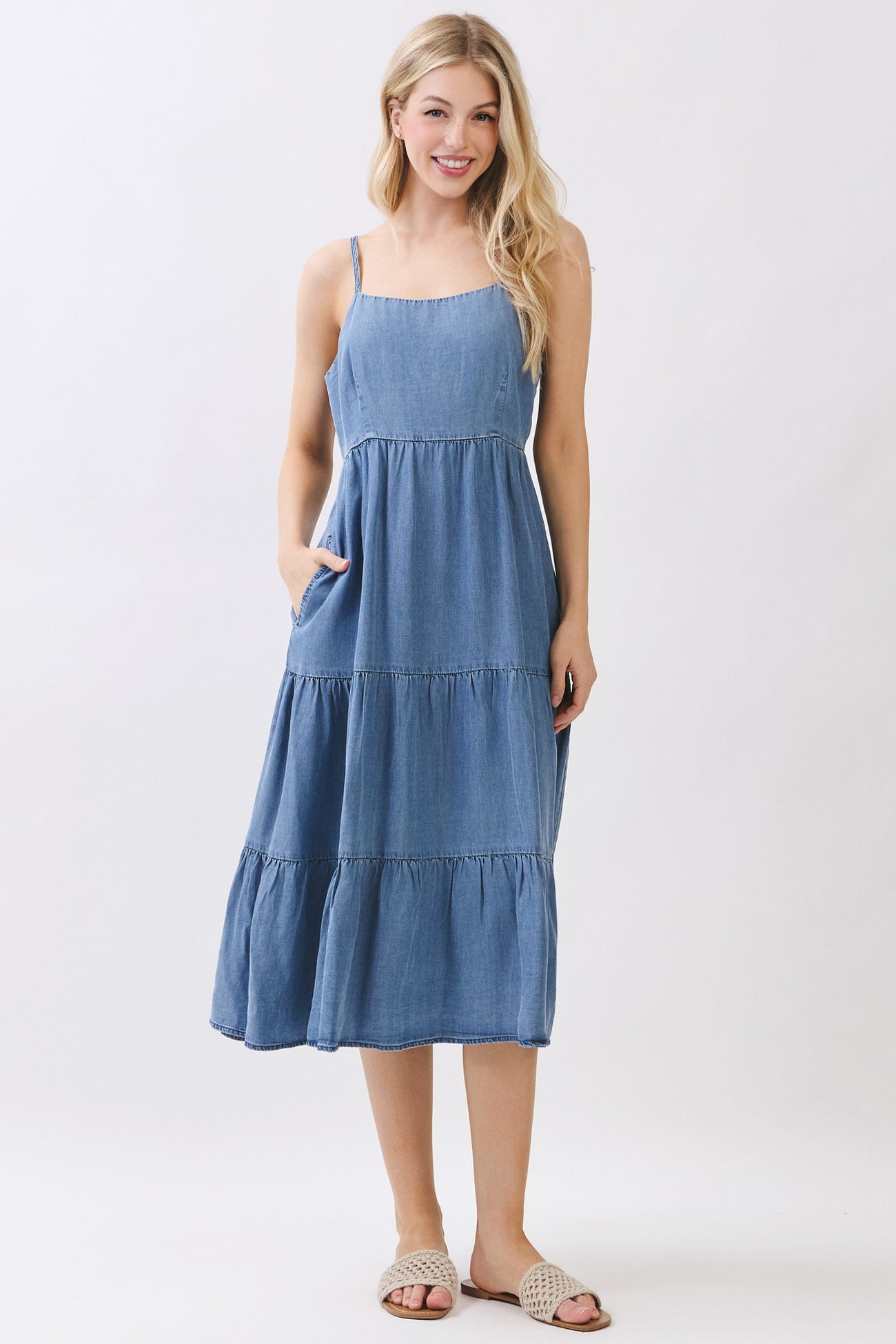 woman wearing a sleeveless denim midi dress with tiered ruffles and pockets, standing and smiling